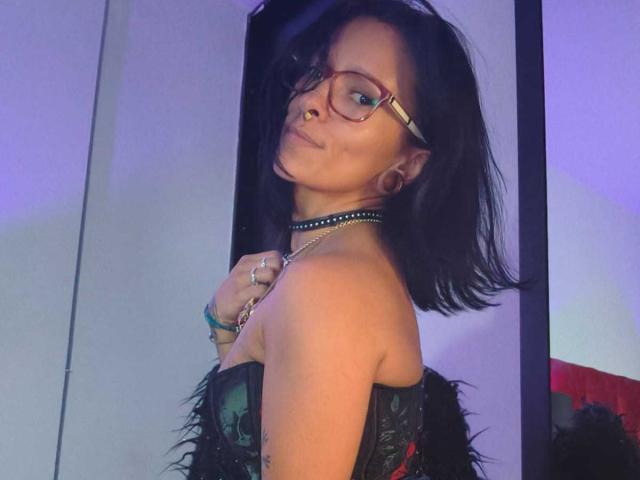 Amelie Blake, met zwart haar, een bril, een neusring en een choker, draagt een zwarte korset met veren, kijkt om, glimlacht en laat haar borsten zien, in een paars verlichte kamer..