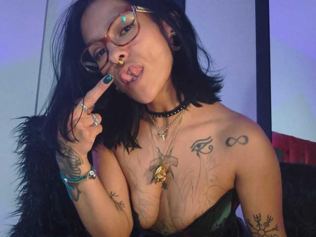 AmelieBlake, a woman with medium brown skin and black shoulder-length hair, wears red glasses and a black choker. She sticks out her tongue and touches her lips with her index finger. She has multiple tattoos, including an infinity symbol on her right shoulder and a floral design on her chest. She wears a strapless green top and has green nail polish. Her right arm has a large tattoo of a bird and a crown. She has a gold ring on her right index finger and a gold pendant necklace. The image is a photograph with a blue and purple light effect.
