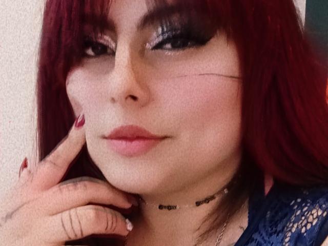 LilithHeavy ha i capelli rossi, l'eyeliner scuro e le labbra rosa, con una piccola cicatrice sulla guancia destra. Indossa una camicetta di pizzo blu e una collana, e sembra soddisfatta..