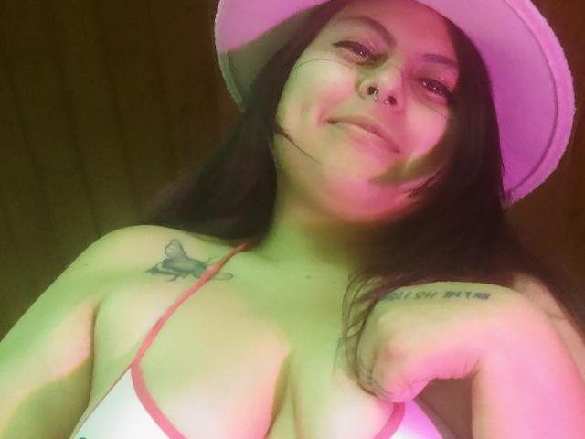 Lilith: Lang zwart haar, een roze hoed, een neusring en tatoeages, toont haar decolleté in een witte top met roze randen, en glimlacht naar de camera..