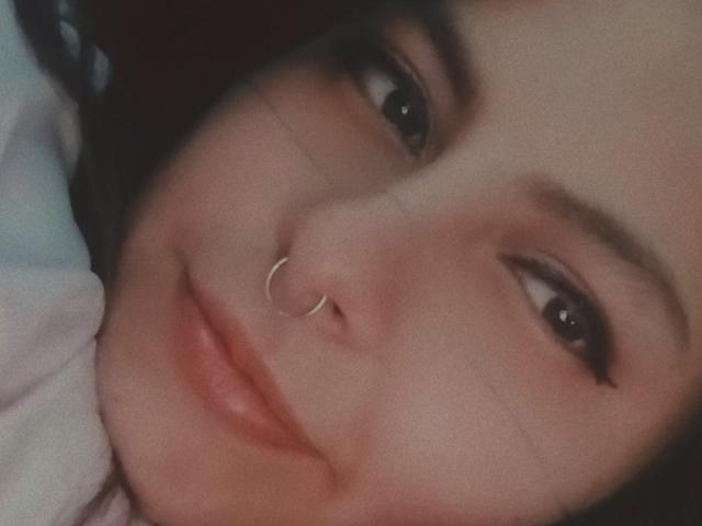 El rostro de LilithHeavy, mostrando una ligera sonrisa con labios rosados y brillantes, cejas oscuras y un piercing de plata en la nariz, en una toma de primer plano con una iluminación suave..