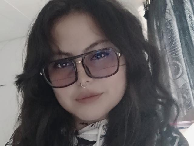 LilithHeavy has long black hair, wears black rectangular glasses, and a nose ring, with blue eyes and a subtle smile, looking slightly to the right.