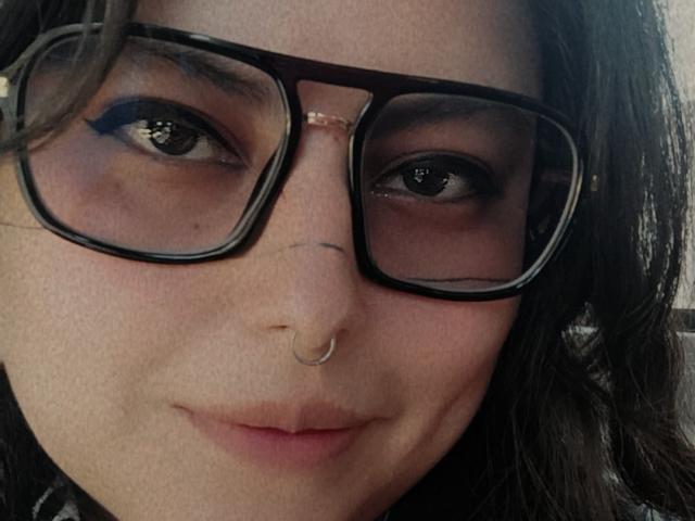 LilithHeavy has dark brown eyes, black-rimmed glasses, and a nose ring. Her skin is light, and she's smiling slightly with dark, wavy hair. Her lips are pink.