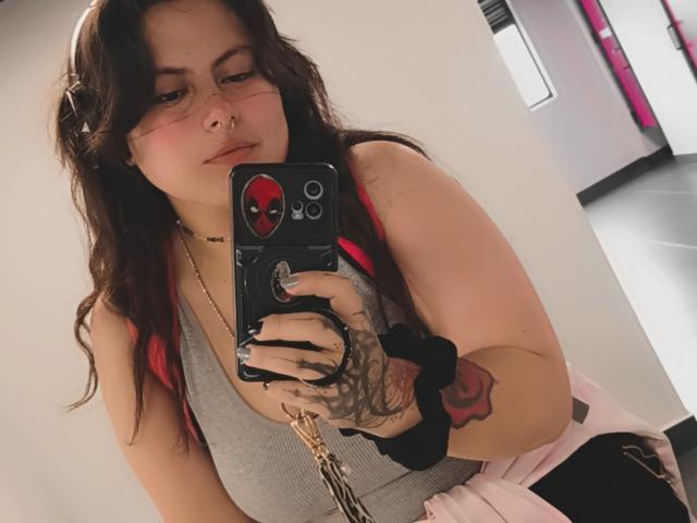 Lilith: Capelli scuri e ondulati, con riflessi rosa, piercing al naso, braccio tatuato, indossa una canotta grigia, una gonna rosa, tiene in mano un telefono con una cover di Spider-Man, selfie allo specchio..