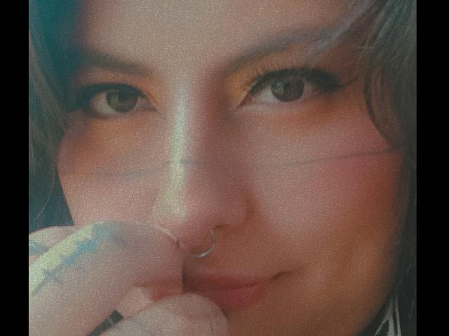 LilithHeavy's face with a nose ring, brown eyes, and a subtle smile, holding her chin with tattooed fingers, soft light, slightly blurry, warm skin tone.