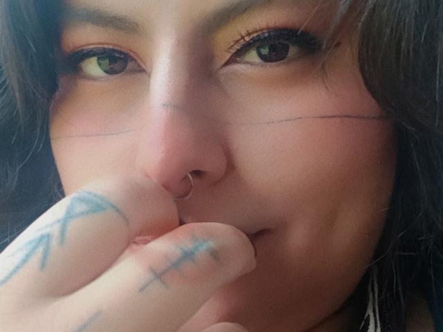 El rostro de LilithHeavy, con el nose piercing, ojos verdes, cabello oscuro, ligeramente sonrojada, tocándose la nariz con dedos tatuados, una sonrisa sutil, piel clara, primer plano..