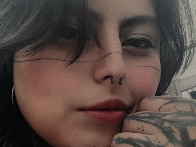 LilithHeavy has a nose piercing, tattooed hand, and dark hair, with subtle makeup and red lips, looking slightly to the side with a relaxed expression.