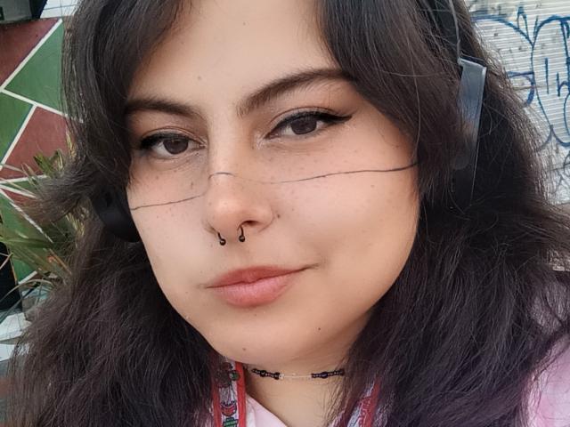 LilithHeavy has dark wavy hair, brown eyes, a nose ring, and black eyeliner with a subtle wing. She's wearing a headset, and her lips are slightly parted.