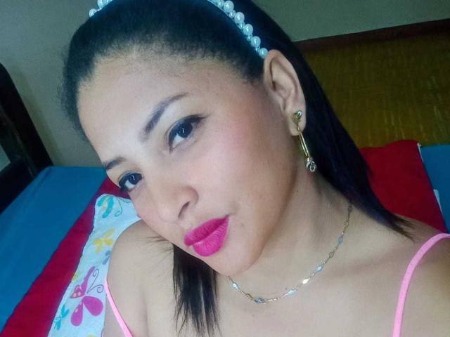 RoxaneMilf, una joven con piel de tono marrón medio y cabello negro lacio, lleva una diadema de perlas y tirantes finos rosas. Su maquillaje incluye delineador de ojos oscuro y un lápiz labial rosa brillante. Tiene aretes de aro dorados y un delicado collar de cadena. Mira ligeramente hacia la izquierda con una expresión neutra. La fotografía es un primer plano, enfocándose en su rostro y hombros superiores. El fondo incluye un cojín azul y rojo con un patrón colorido. La imagen tiene un aspecto casual y espontáneo..