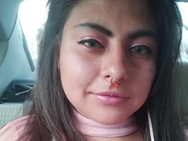LilithHeavy has pink glitter eyeshadow, a red nose ring with small red jewels, pink lipstick, and long brown hair. She's wearing a pink choker and a pink shirt.