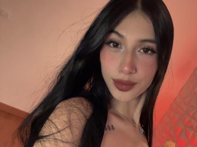 SophieBeaumont with long black hair, brown eyes, dark lipstick, nose ring, and tattoo on her chest, looking at the camera with a slight smile. Her tits are partially visible.