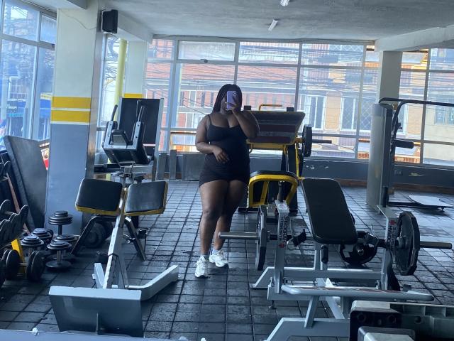 A gym photo shows Dilay, a dark-skinned woman with a curvy physique, standing in the center. She wears a black strapless mini dress and white sneakers. Dilay holds a smartphone in front of her face, partially covering it. The gym is filled with workout equipment, including a yellow weight machine and black benches. The floor is tiled with dark squares. Natural light comes through large windows behind her, revealing a building exterior. Dilay's hair is dark, and she wears a colorful face mask.