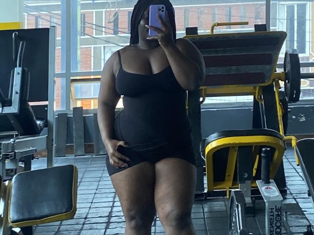 Dilay, dark-skinned, curvy with large tits and ass, wearing a black tank top and shorts, taking a mirror selfie in a gym, holding a purple phone, standing.