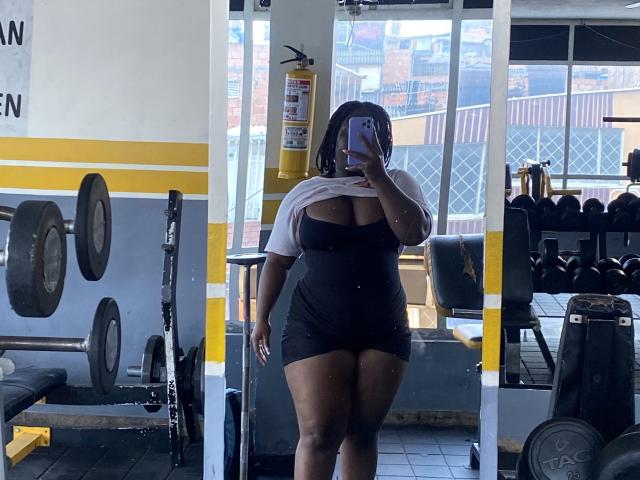 Dilay stands in a gym, taking a mirror selfie. She's a curvy woman with dark skin and shoulder-length hair, wearing a white crop top and black mini dress. Her right arm is lifted, revealing her cleavage. The gym has weight racks, dumbbells, and large windows with cityscape views. Dilay holds a smartphone in her left hand. The photo has natural light, emphasizing her figure and the gym's equipment.