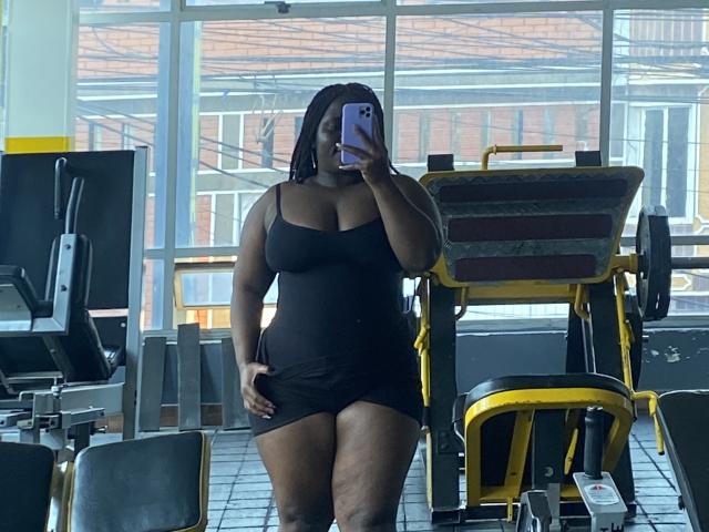 A photo of Dilay in a gym. She's standing in the center, taking a mirror selfie with a purple phone. She's wearing a tight black dress that accentuates her curvy, plus-sized physique. Her dark skin contrasts with the black dress. She has shoulder-length black hair. The gym has yellow and gray equipment in the background, and large windows letting in daylight. Dilay's right hand rests on her thigh. The image has a casual, realistic style.