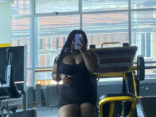 Dilay with big tits and dark skin, wearing a black tank top and shorts, taking a mirror selfie in a gym with long braids, holding a purple phone.