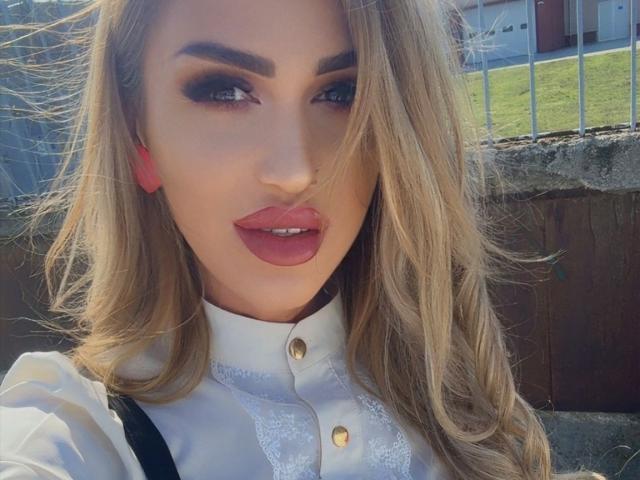 NicolSNaya has blonde hair, blue eyes, dark eyeliner, pink lips, and wears a white button-up shirt with lace. Her pink earrings stand out against her fair skin.