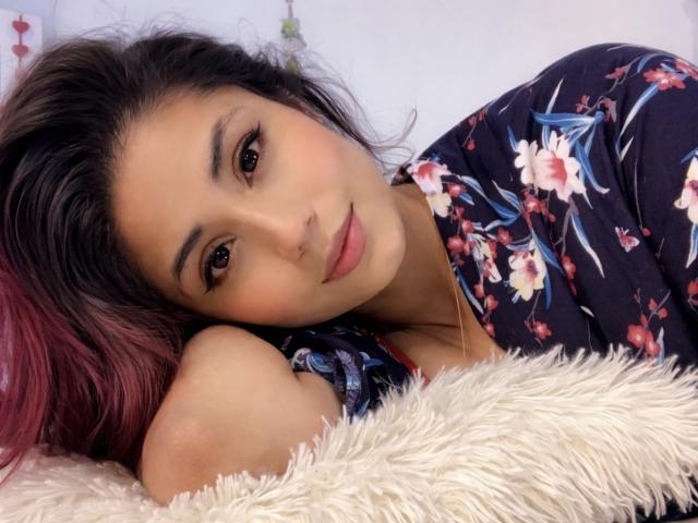 KattyWattson with red-pink ombre hair, floral dress, lying on fluffy white pillow, head resting on arm, brown eyes, subtle makeup, soft smile, dark eyebrows, cute face.