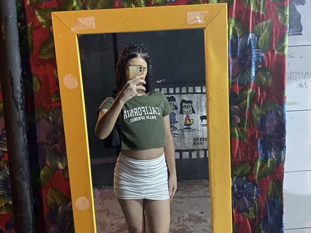 Gabrielly69 stands in front of a mirror with a yellow frame, wearing a green crop top and white ruffled skirt, holding a phone, showing her small ass and tits.