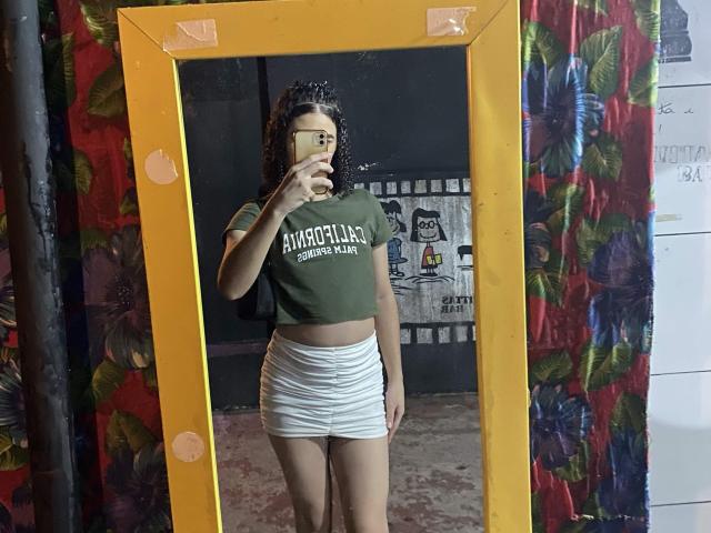 Gabrielly69 with curly hair, glasses, green crop top, white ruched skirt, taking a mirror selfie, yellow frame, colorful floral curtain, holding phone, standing in room.
