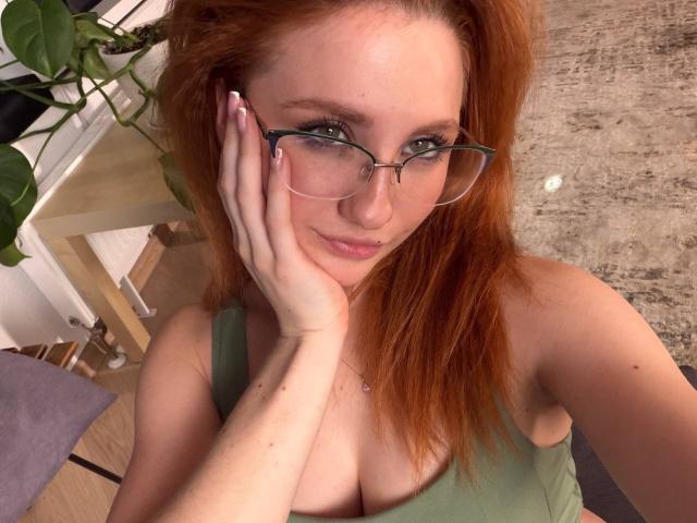 JenniferOhWow with red hair, green tank top, glasses, and hand on cheek, showing her tits. She's smiling, looking up, with a necklace. Her nails are manicured.