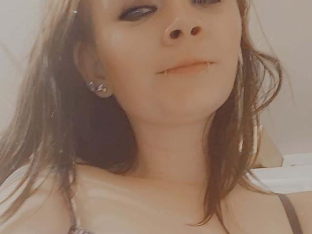 SpreadFun with brown hair, pale skin, black eyeliner, small nose, and lip piercing, wearing a black strap, slightly smiling, showing collarbone and upper chest.