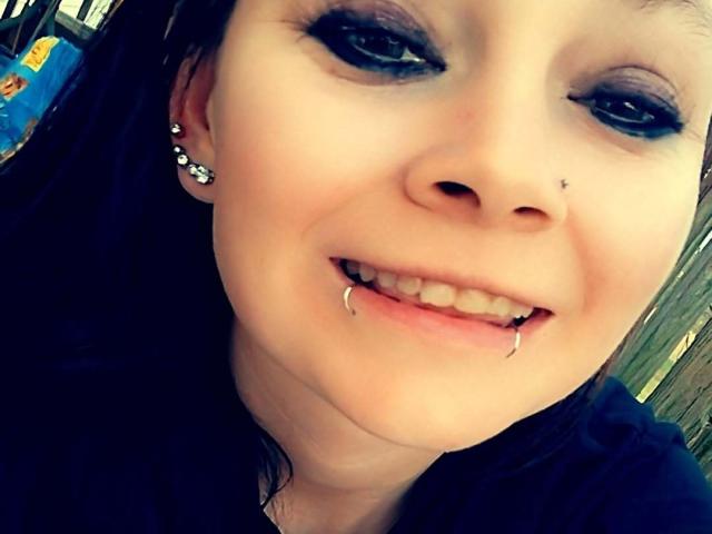 SpreadFun has pale skin, dark eyeliner, a nose piercing, and a lower lip piercing with fang-like jewelry. She's smiling with blue eyes and black hair.