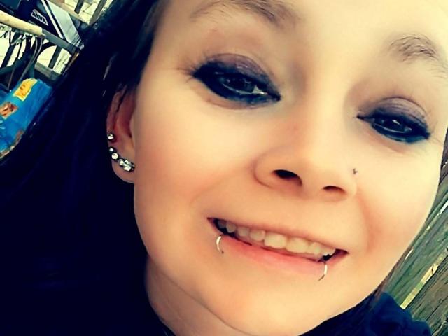 SpreadFun has a pale face with dark blue eyeliner, pierced ears, a nose stud, and a lower lip piercing with small metal balls. She's smiling with her teeth slightly visible.