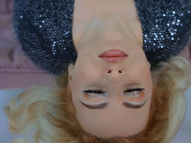 JenniferJane's face is upside down, with eyes closed, wearing shiny silver sequins, pink lipstick, and blonde hair, lying on a pink surface. Her tits are partially visible.