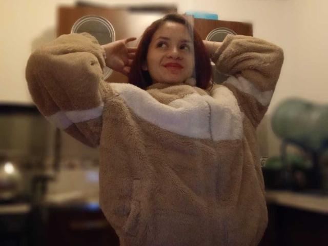 LesleyGray with red hair, smiling, wearing a fluffy brown and white fleece jacket, hands behind head, standing in a kitchen, slightly blurred. Boobs not visible.