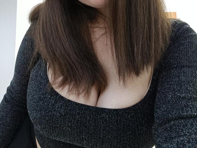 Jaklinna's big tits in a black, textured, low-cut top with her brown hair covering her chest slightly. Her pale skin is visible between the top and hair.