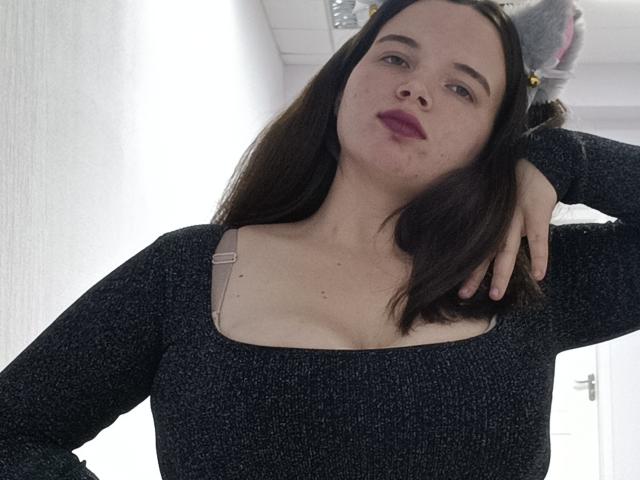 Jaklinna with dark brown hair, wearing a black textured sweater, exposing small tits through a square neckline, hand on head, pink lipstick, white background, and a fluffy gray cat.