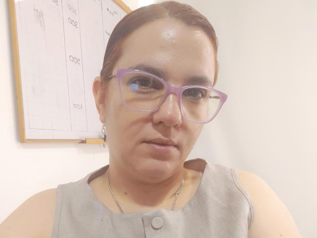 AmeliaWilson has a chubby face, brown skin, and is wearing purple glasses. She's dressed in a gray sleeveless top with a button and a silver necklace.