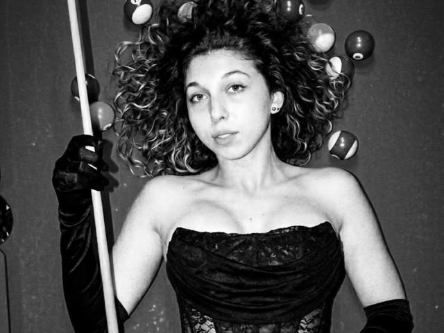 Black-and-white photo of Sanella with curly, voluminous hair. She wears a strapless, lace-patterned top and long, black gloves. Sanella holds a pole with her right hand, looking directly at the camera with a neutral expression. Balloons float around her head. Her light skin contrasts with the dark lace and gloves. Her earrings are small and round. The image has a classic, 20th-century style.