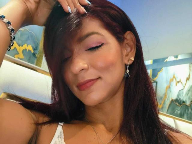HannaPretty has dark red hair, slightly parted to the side, with her right hand touching her hair. She has pink eyeliner, a subtle pink blush, and red lipstick. She wears small, gold earrings and a white lace top. Her left wrist has a blue beaded bracelet. The background is blurred, with abstract blue and yellow patterns. Her skin is warm-toned, and she has a slight smile. The image has a soft, warm light.