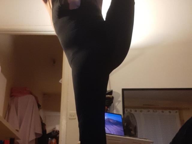 Close-up of IssahneMay's right leg, bent at the knee, wearing tight black leggings. The leg is centered in the frame, highlighting the stretch and the fabric's texture. The background includes a partially open door, a pink garment on a shelf, and a TV with a blue screen. Warm, indoor lighting. The focus is on the leg's form and the leggings' material. The image has a casual, slightly cluttered home setting.