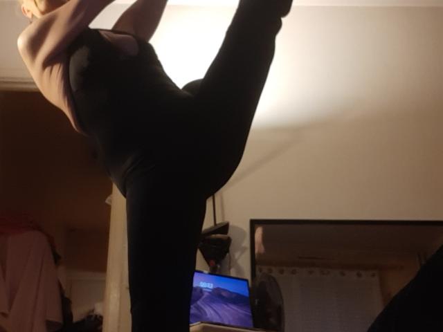 IssahneMay is doing a high kick in black pants and a black top, showing her bare tits and her ass is raised. She's in a dimly lit room.