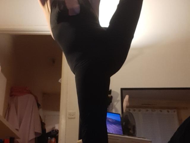 Close-up of IssahneMay's black, form-fitting leggings with a raised leg, focusing on the thigh and hip area. Her hand is near the top, partially visible. The room has a mirror reflecting part of her lower body. Background includes a pink shirt on a shelf, a white wall, and a computer screen showing a blue landscape. The image is slightly blurred, emphasizing the leg and hand.