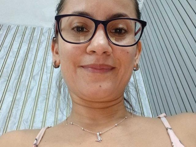 SolaraDubois, a medium-skinned woman with dark brown hair in a ponytail, wears black-framed glasses, small hoop earrings, and a pink strap dress. She has a small mole on her left cheek and a subtle smile. She's looking directly at the camera. The background features a striped pillow and a striped wall. She wears a silver necklace with a small pendant. The image is a close-up selfie.