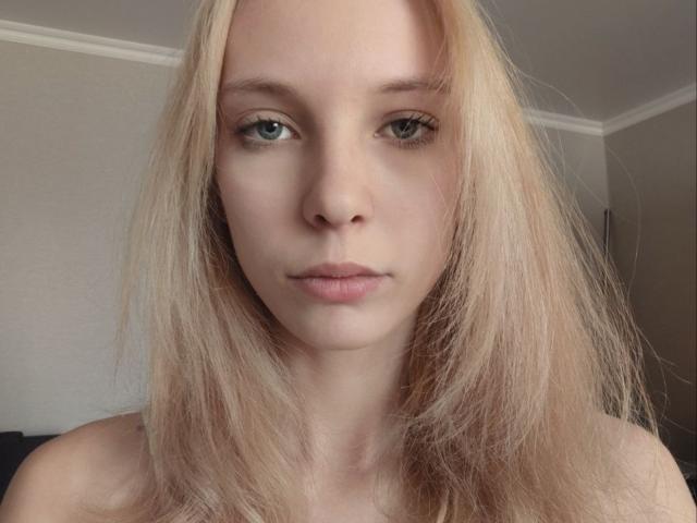 LillianRouselle has blonde hair, green eyes, and a neutral expression. Her bare shoulders are visible. She's staring straight at the camera with a slightly pouty, relaxed face.