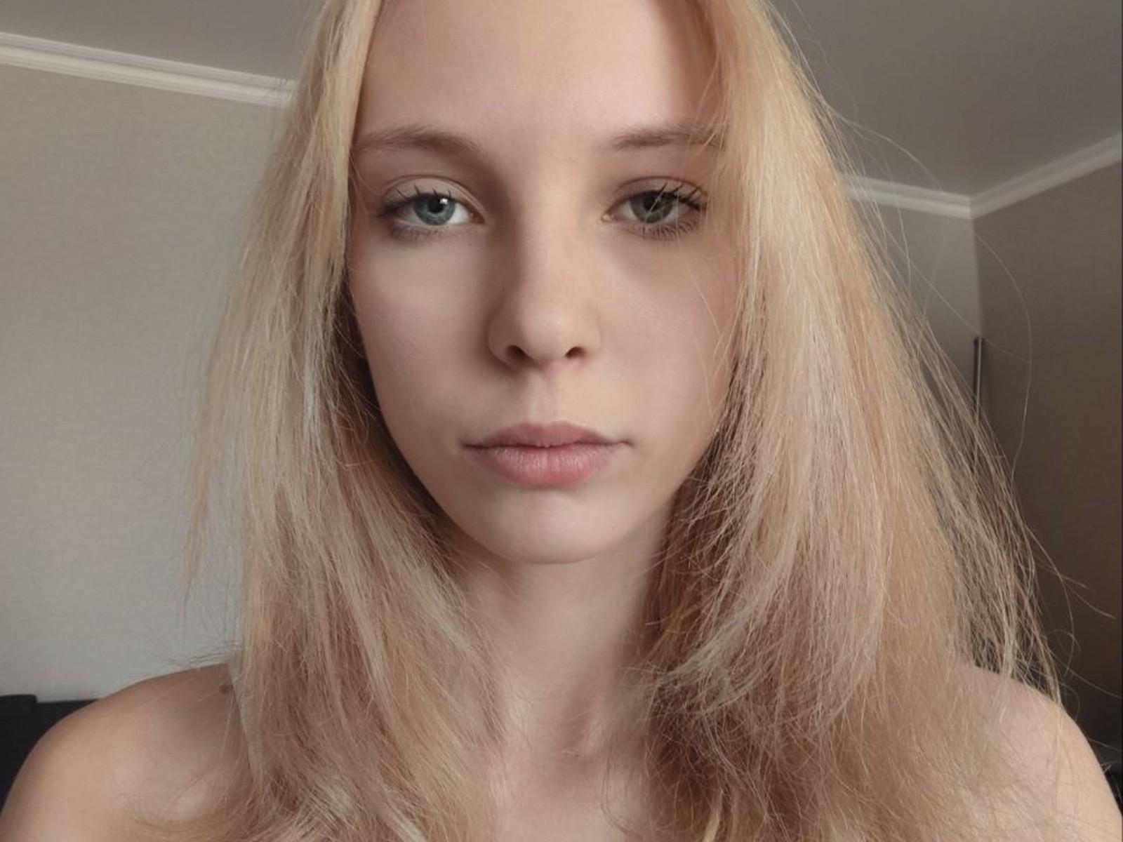 LillianRouselle has blonde hair, green eyes, and a neutral expression. Her bare shoulders are visible. She's staring straight at the camera with a slightly pouty, relaxed face.