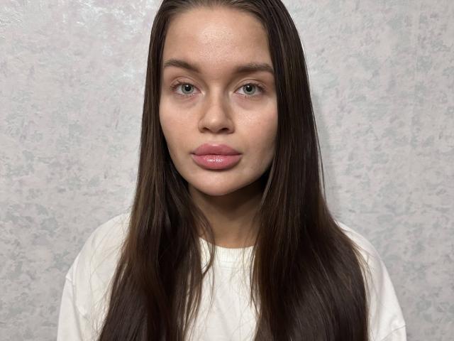IngaSelander has long brown hair, light skin, green eyes, and pink glossy lips, wearing a white shirt, with a subtle, textured background. Her face is centered in the photo.