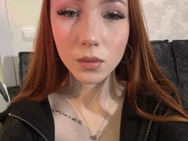 AlonaHadcock has red hair, fair skin, and is wearing a black zip-up. She has a tattoo under her chin, hoop earrings, and is looking at the camera with pouty lips.
