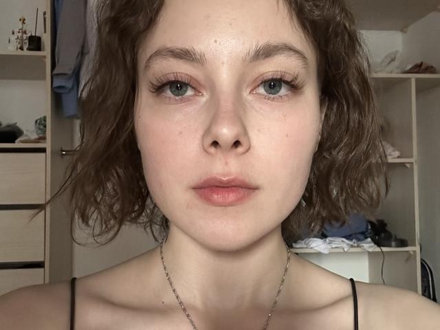 KristalRozon has curly brown hair, pale skin, blue eyes with long eyelashes, and pink lips. She's wearing a black spaghetti strap top and a silver chain necklace.