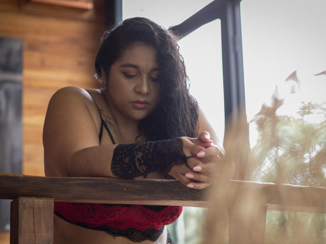 ViickyLee, a young woman with medium brown skin and wavy black hair, leans over a wooden railing. She wears a black lace glove on her right hand and a red lace bra. Her eyes are downcast, and her expression is serious. The wooden structure and blurred outdoor elements are in the background. Soft natural light highlights her features and the lace textures. The image has a warm, slightly muted color palette.