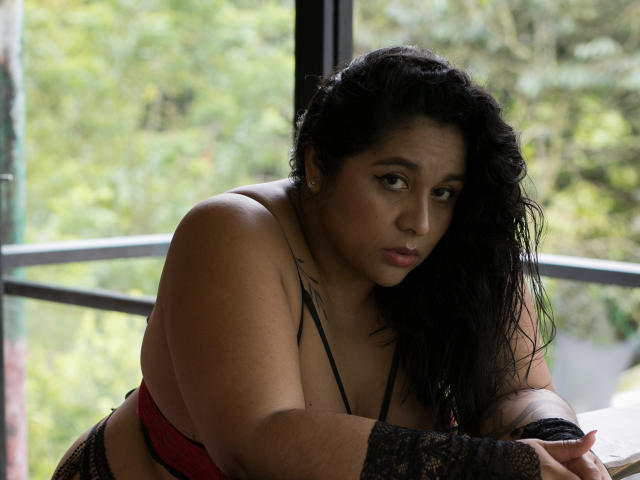 ViickyLee, a plus-sized woman with dark, wavy hair, sits in a relaxed pose. She wears a black lace sleeve and red bra, looking slightly to the side with a serious expression. Her tattoo on the left shoulder is partially visible. The light is soft, highlighting her smooth, tan skin. The background is blurred greenery through a window. The image has a natural, intimate feel.