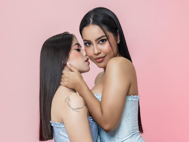 Two women with long, straight black hair stand close, facing each other. PaolaYKatty, on the right, has tan skin and wears a light blue, strapless, ruffled corset. She gently holds the chin of the other woman, who has fair skin and a small tattoo on her upper arm. Both women have subtle makeup with dark eyeliner and pink lipstick. PaolaYKatty's expression is soft, while the other woman looks at her with a slight smile. The light blue corset contrasts with their skin tones. The composition focuses on their faces and upper bodies.