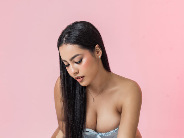 PaolaYKatty has long, straight black hair, slightly parted to the left. She's looking down with a subtle, thoughtful expression. Her skin is smooth and tan, with pinkish blush on her cheeks. She's wearing a strapless blue-gray top that reveals her large, natural breasts. She has small, gold earrings and a delicate necklace with a small cross. The background is a solid pink. The lighting is soft, highlighting her features and the texture of her hair. She has full, dark eyebrows and dark eyeliner. The overall composition is centered and focuses on her upper body and face.