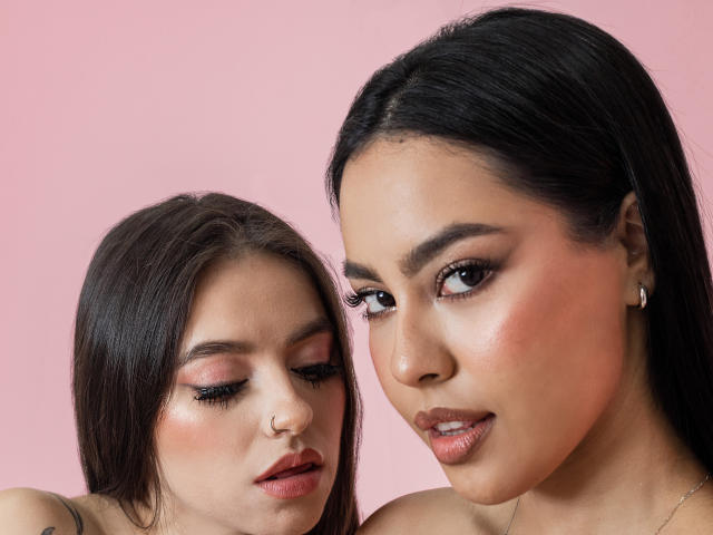 Close-up of two women against a pink background. PaolaYKatty on the right has dark hair, brown eyes, and subtle pink blush. She wears small earrings and has a slight smile. The woman on the left has lighter brown hair, closed eyes, and a nose ring. Both have shimmering eyeshadow and long eyelashes. The right woman's skin is slightly darker than the left woman's. The image uses soft lighting and a focus on facial features.
