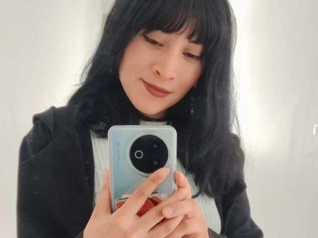 IssisDiosa, avec des cheveux noirs et une frange, tenant un téléphone avec une coque bleue, souriant légèrement, portant une veste noire par-dessus une chemise blanche, dans un autoportrait..